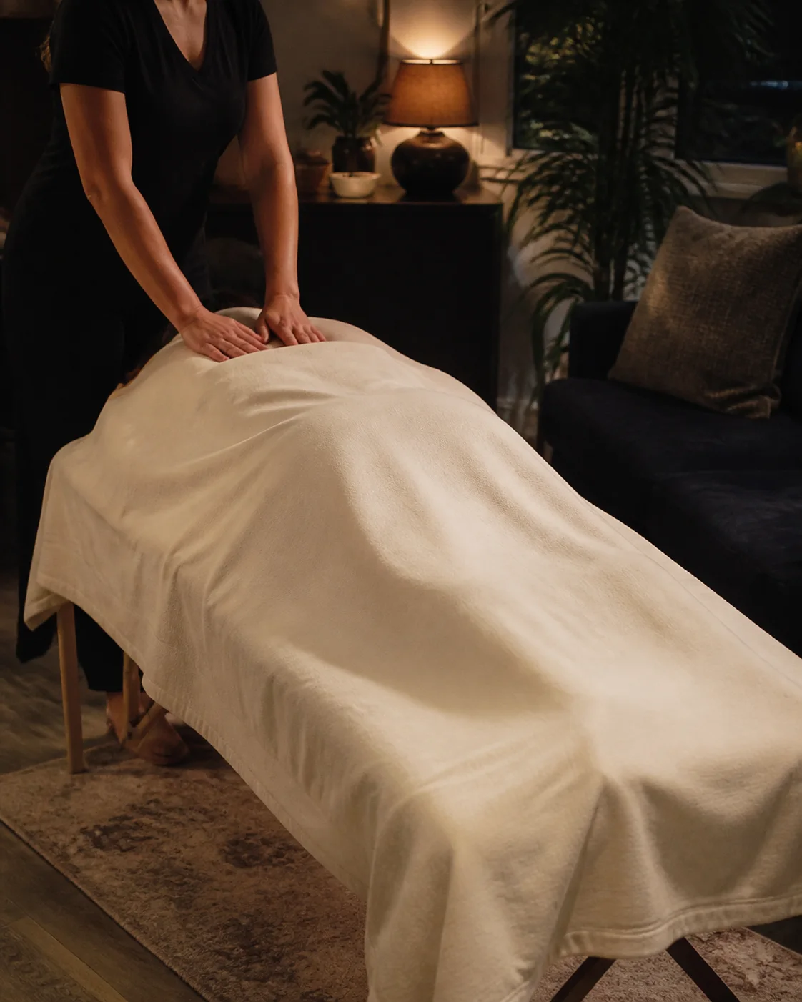 Therapist applying steady pressure during a massage on a draped table in a warmly lit living room.